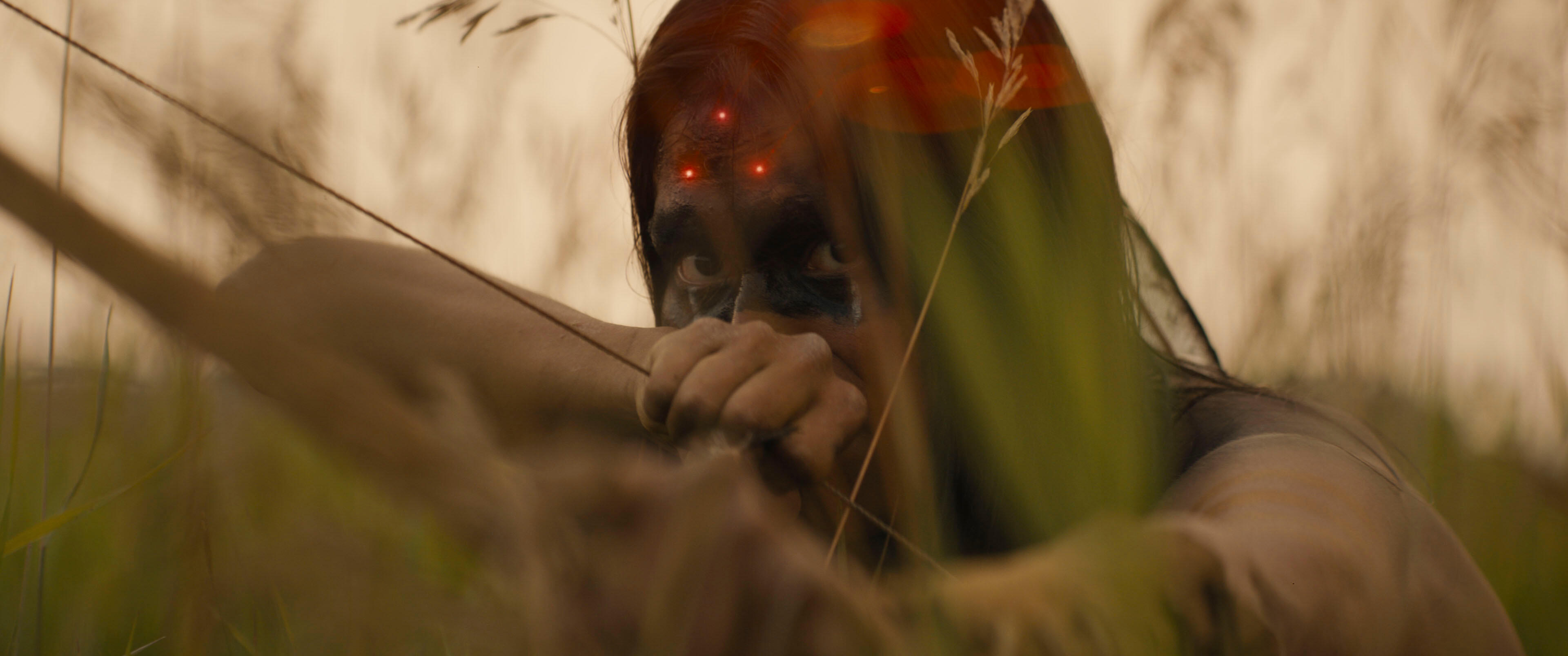 Itsee (Harlan Kytwayhat) draws their bow as a three-pronged laser sight is visible on their forehead in Prey.